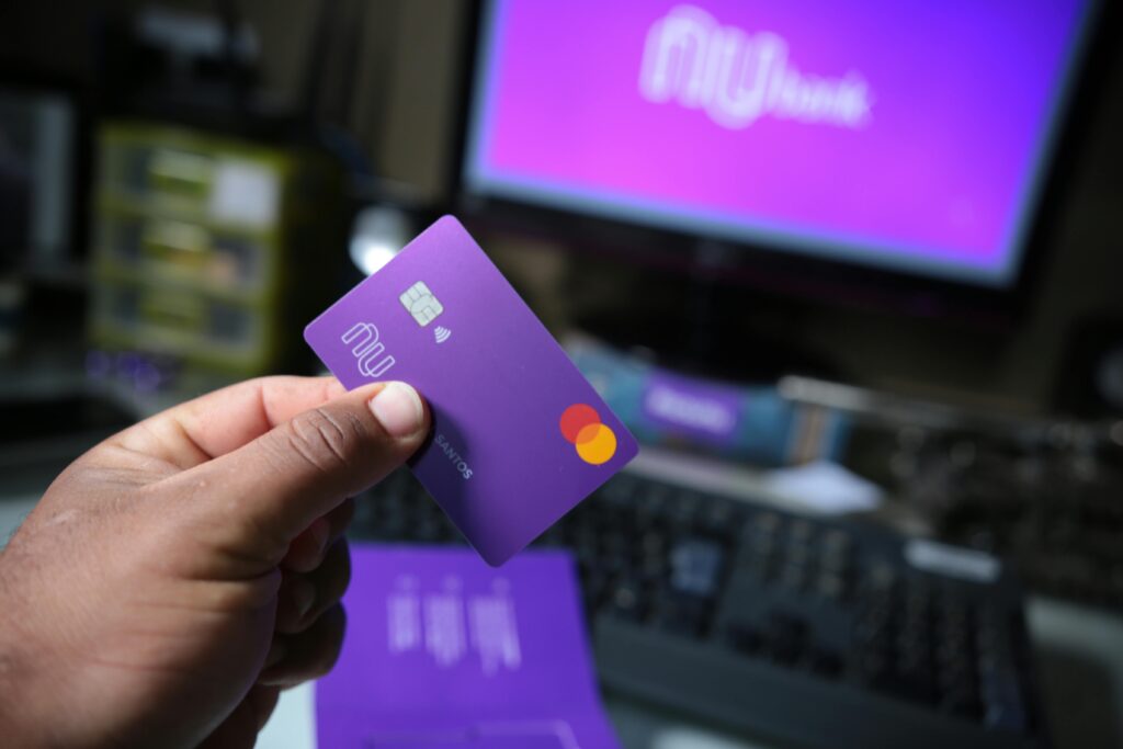 Close-up of a hand holding a purple bank card with a computer setup in the background, signifying online transactions.
